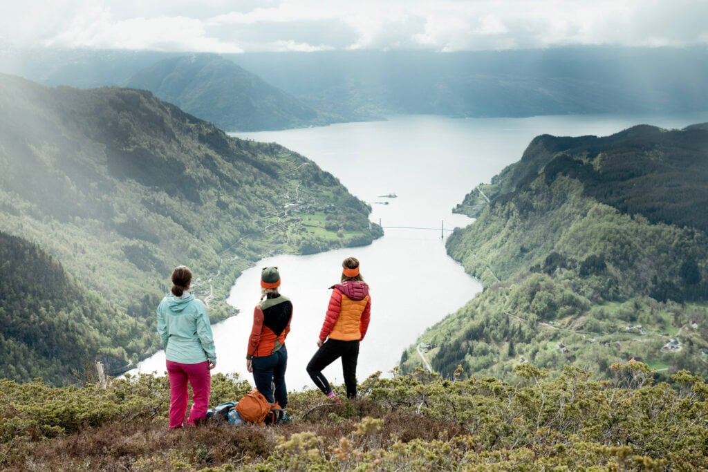 Three woman on a mountain slope looking down at the fjord and bridge below them.