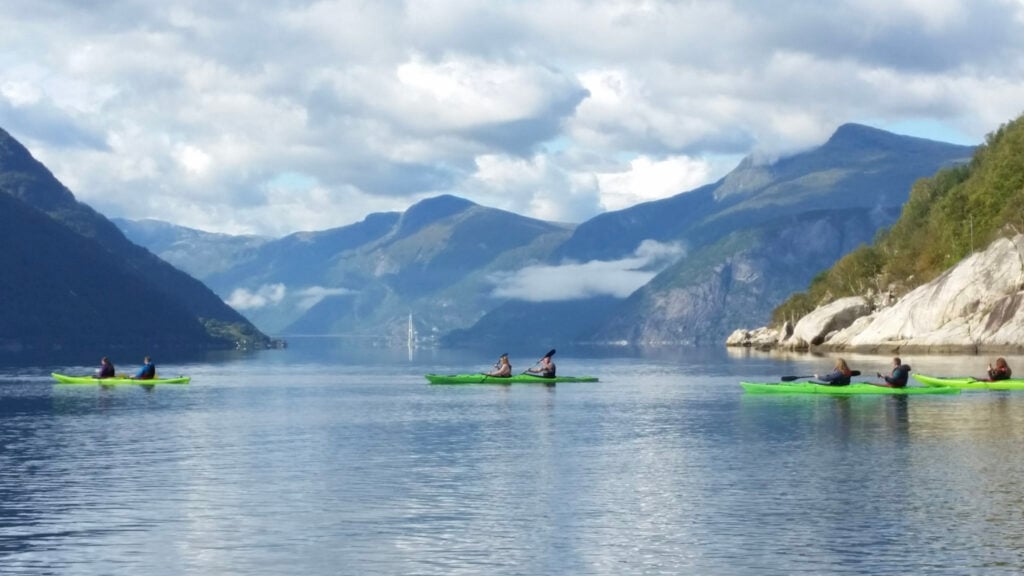 Four green double kayaks on a fjord with mountains and a bridge in the background.