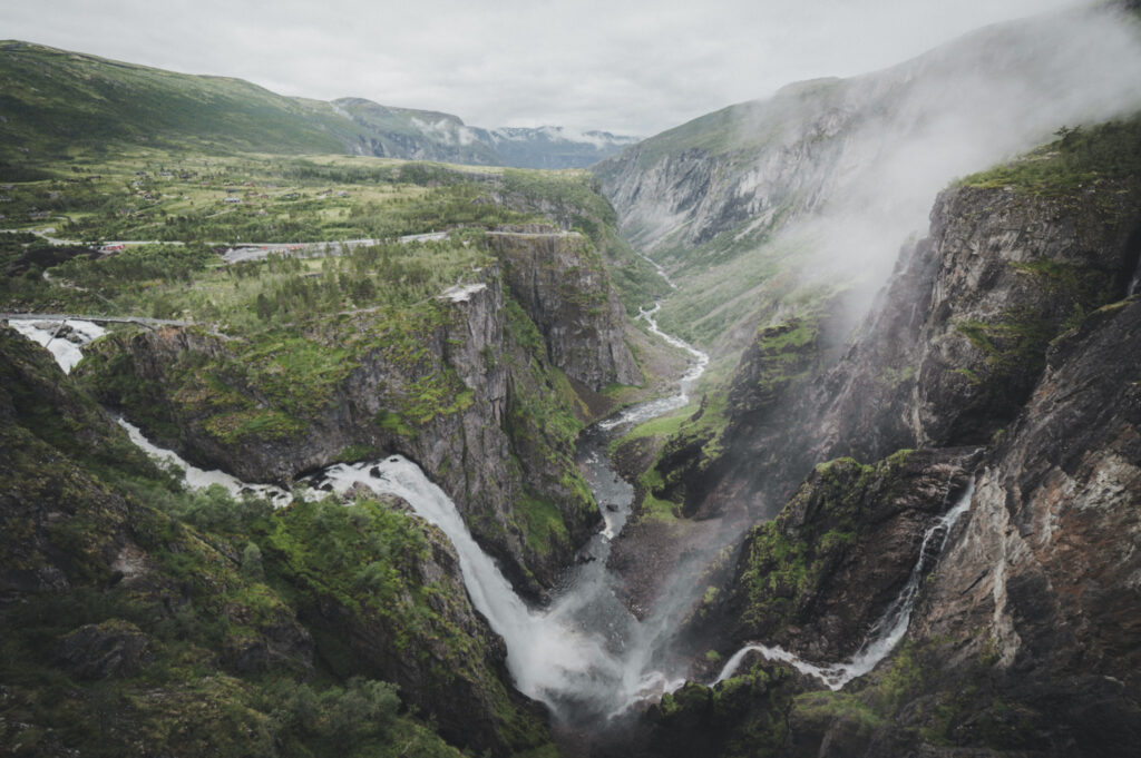 Wasserfall mit Ästen auf beiden Seiten, der in ein Tal hinabstürzt, in dessen Mitte ein Fluss verläuft.