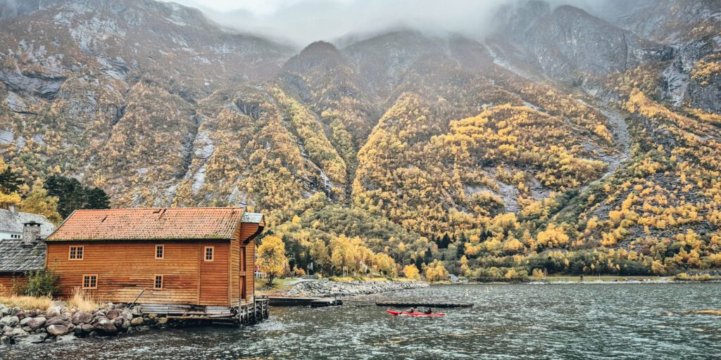 A tandem kayaking paddling into shore by a yellow boat house in Eidfjord. Autumn coloured mountains partly covered by fog.