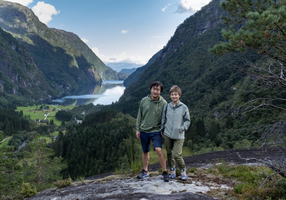 Two people posing on top of a mountain with a fjord in the background.
