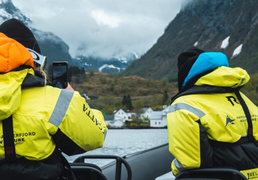 Two people in yellow floatation suits on a boat, one of whom is taking a picture with a mobile phone.